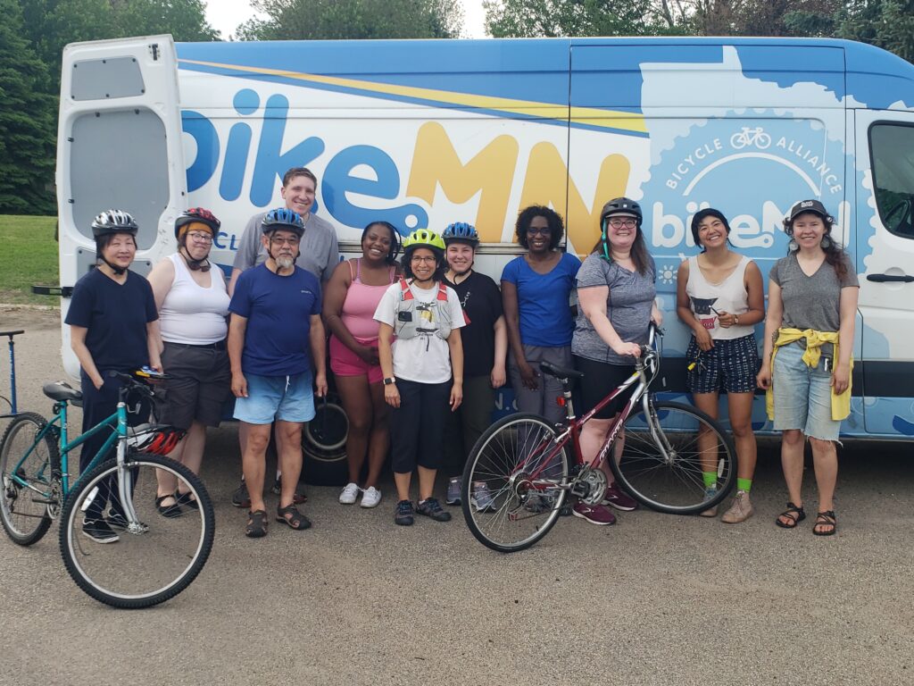 About 12 people stand outdoors in front of the BikeMN van, some have helmets on and their bikes with them. 