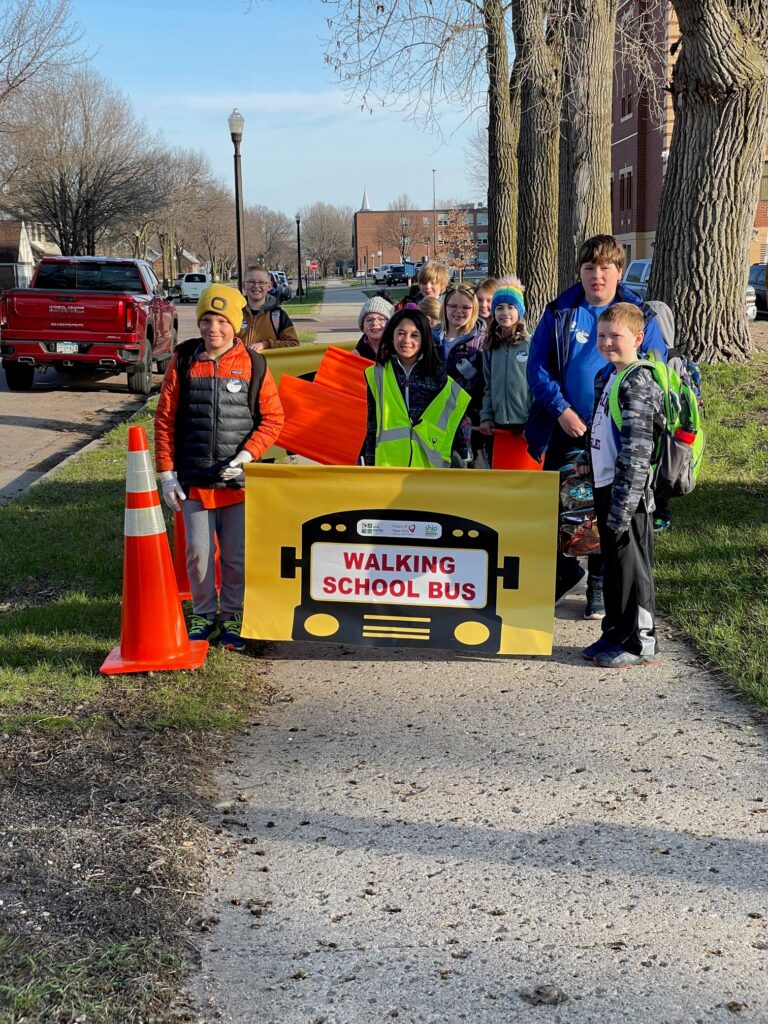 A walking school bus in New Ulm, Minnesota. Students, families, and educators gather behind a yellow walking school bus sign on a path outdoors.