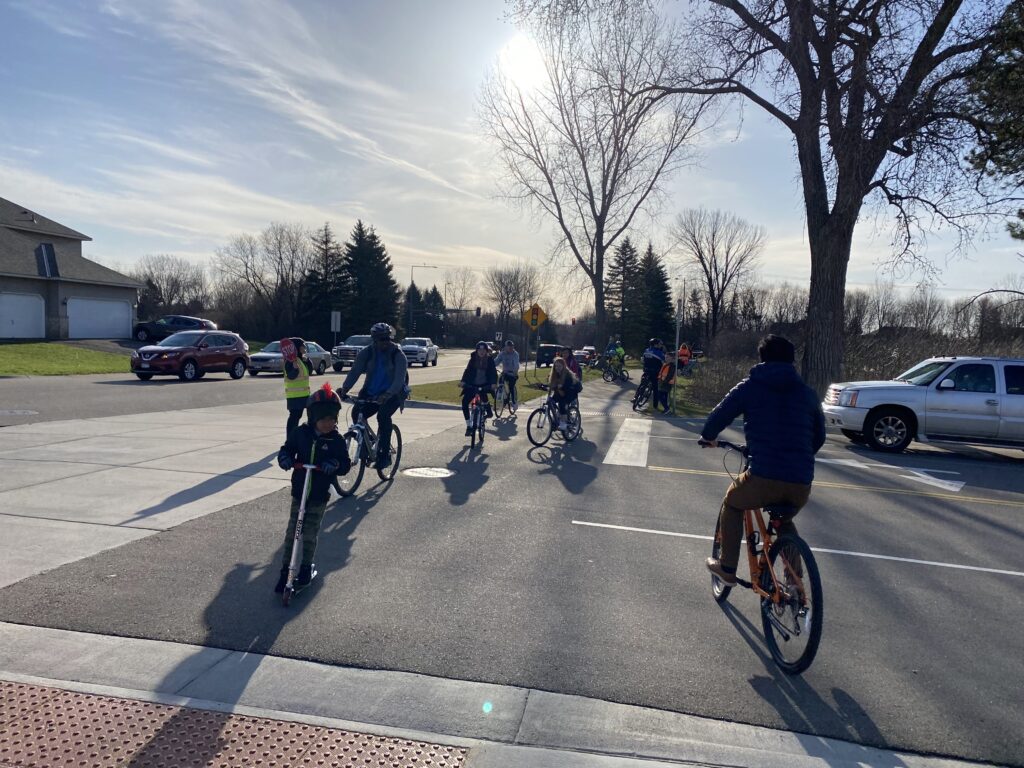 People riding bikes and scooters safely cross the street with the help of a crossing guard. 