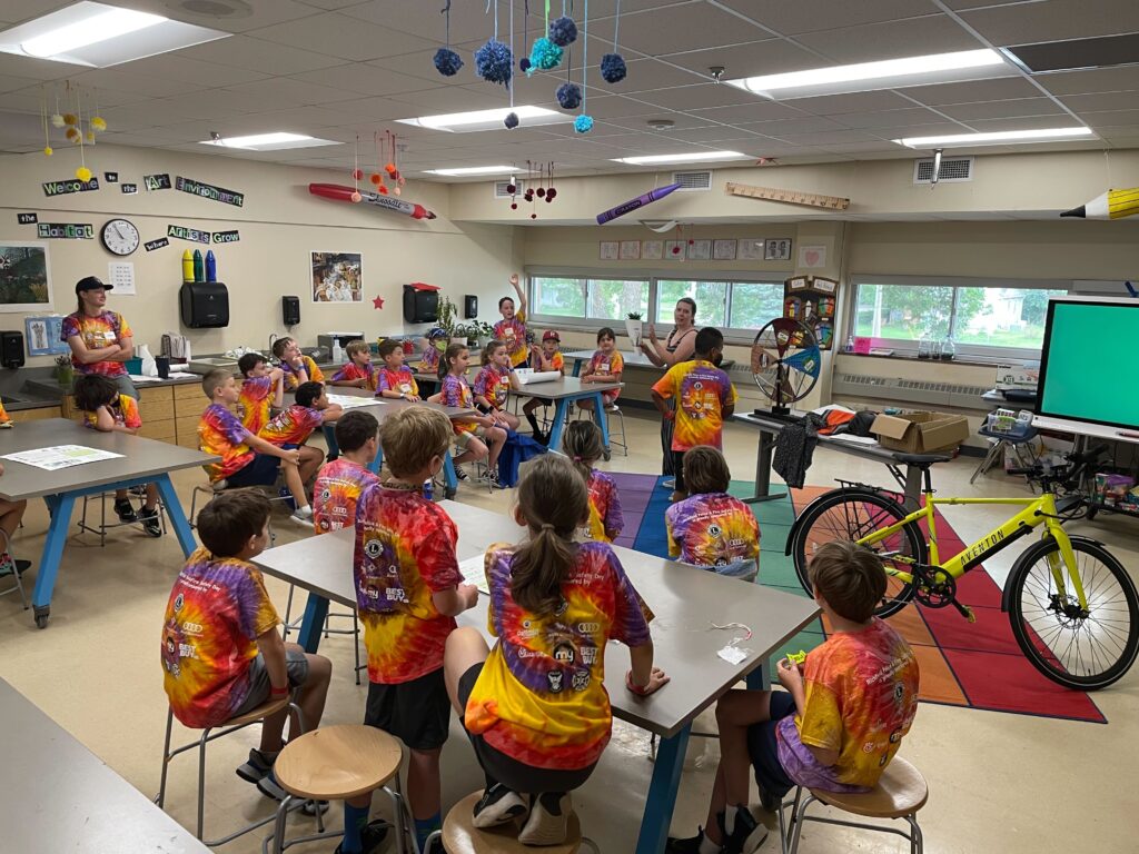 Over fifteen students sit on tables or on the floor and listen to Angela, who is standing at the front of the classroom. A bright yellow bike is on the right side of the photo. 